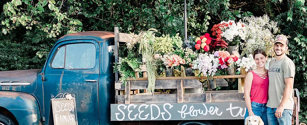 Two people standing with their dog in front of a blue vintage farm truck that has a beautiful flower display in the back