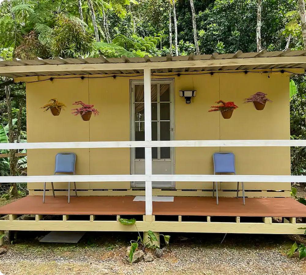 Featured Image - yellow cabin with hanging plants and two blue chairs