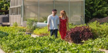 a couple walking through a garden with a greenhouse in the background